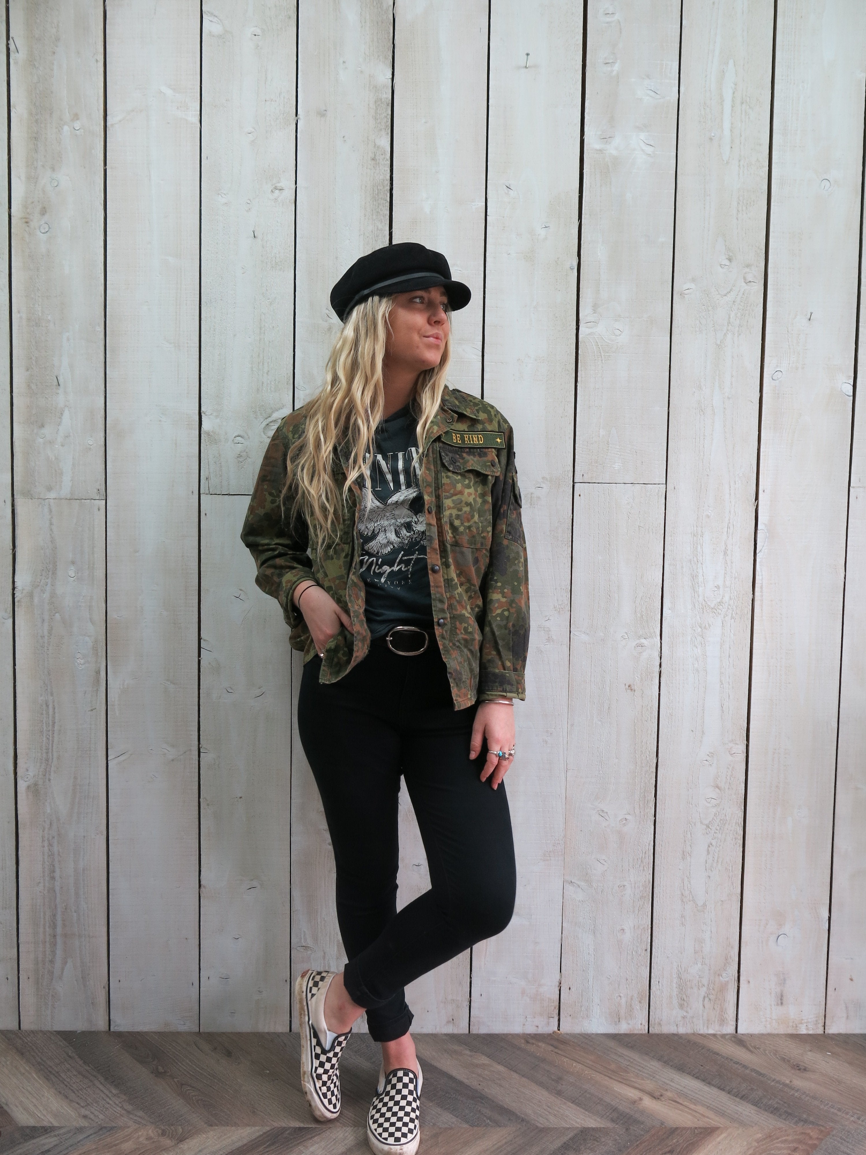 Person wearing a camouflage military jacket with a "BE KIND" embroidered chest patch and military-inspired sleeve patches.