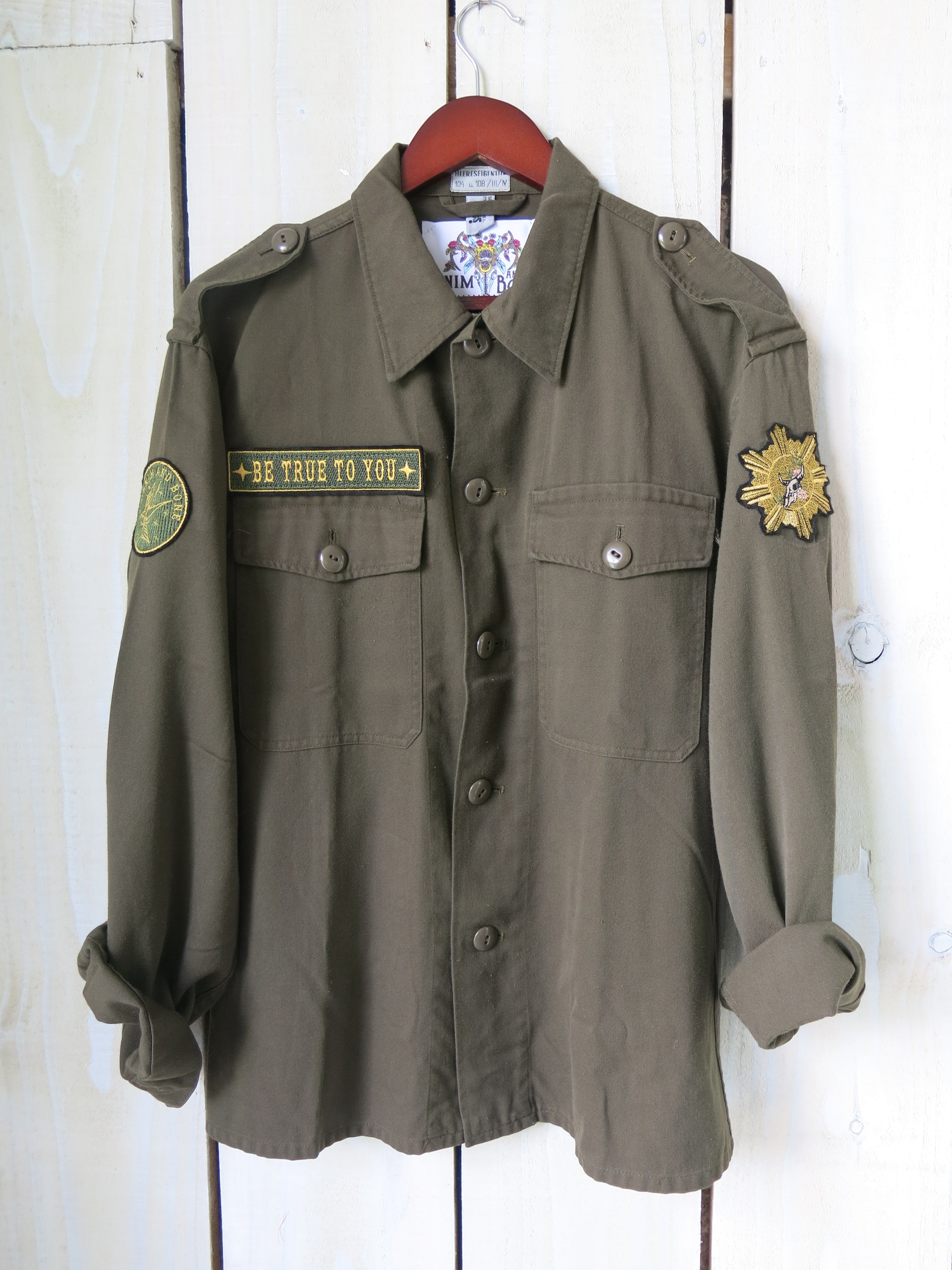 Front view of an olive green military jacket with a "BE TRUE TO YOU" embroidered chest patch and military-inspired sleeve patches.