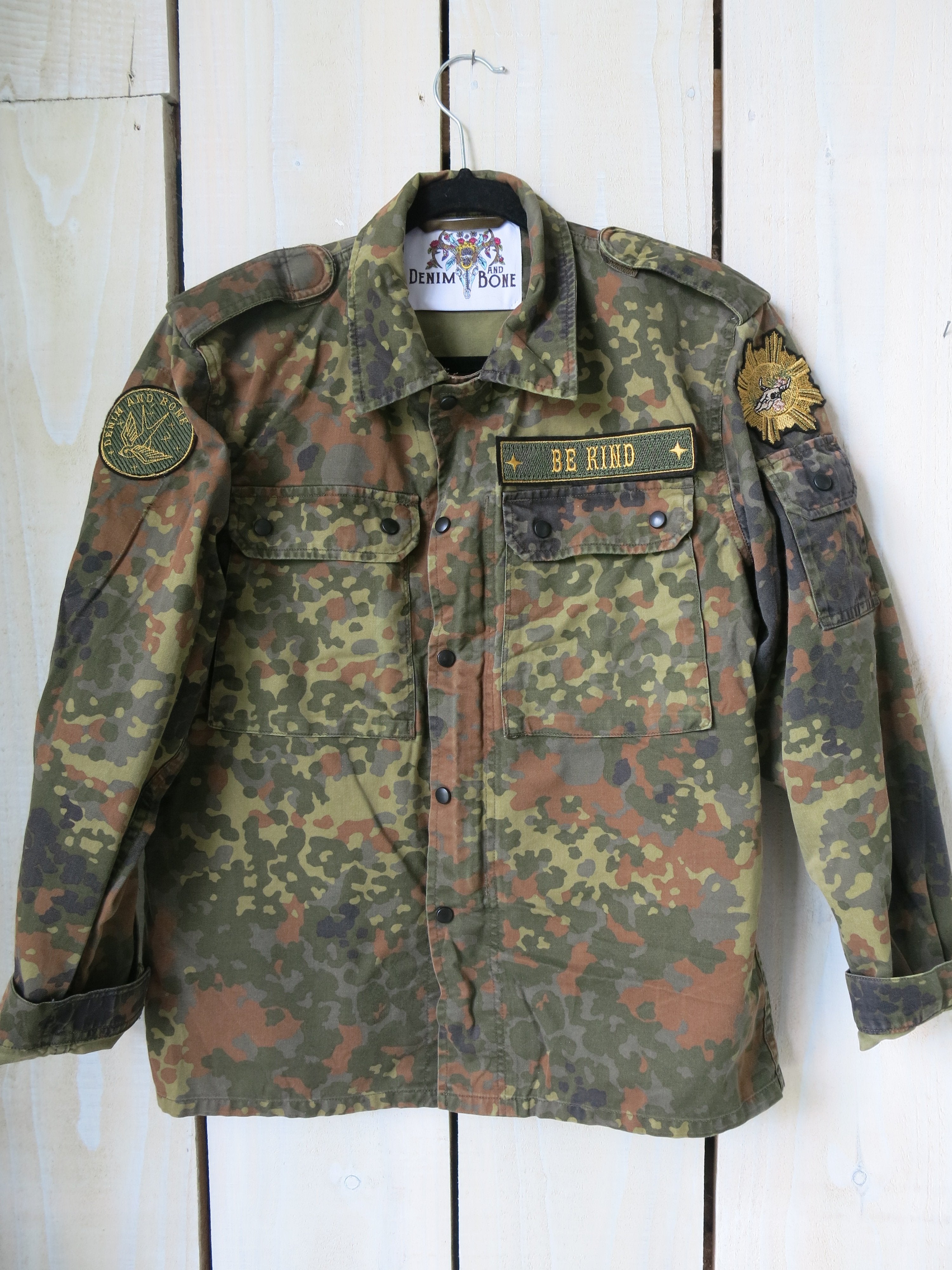 Front view of a camouflage military jacket with a "BE KIND" embroidered chest patch and military-inspired sleeve patches.
