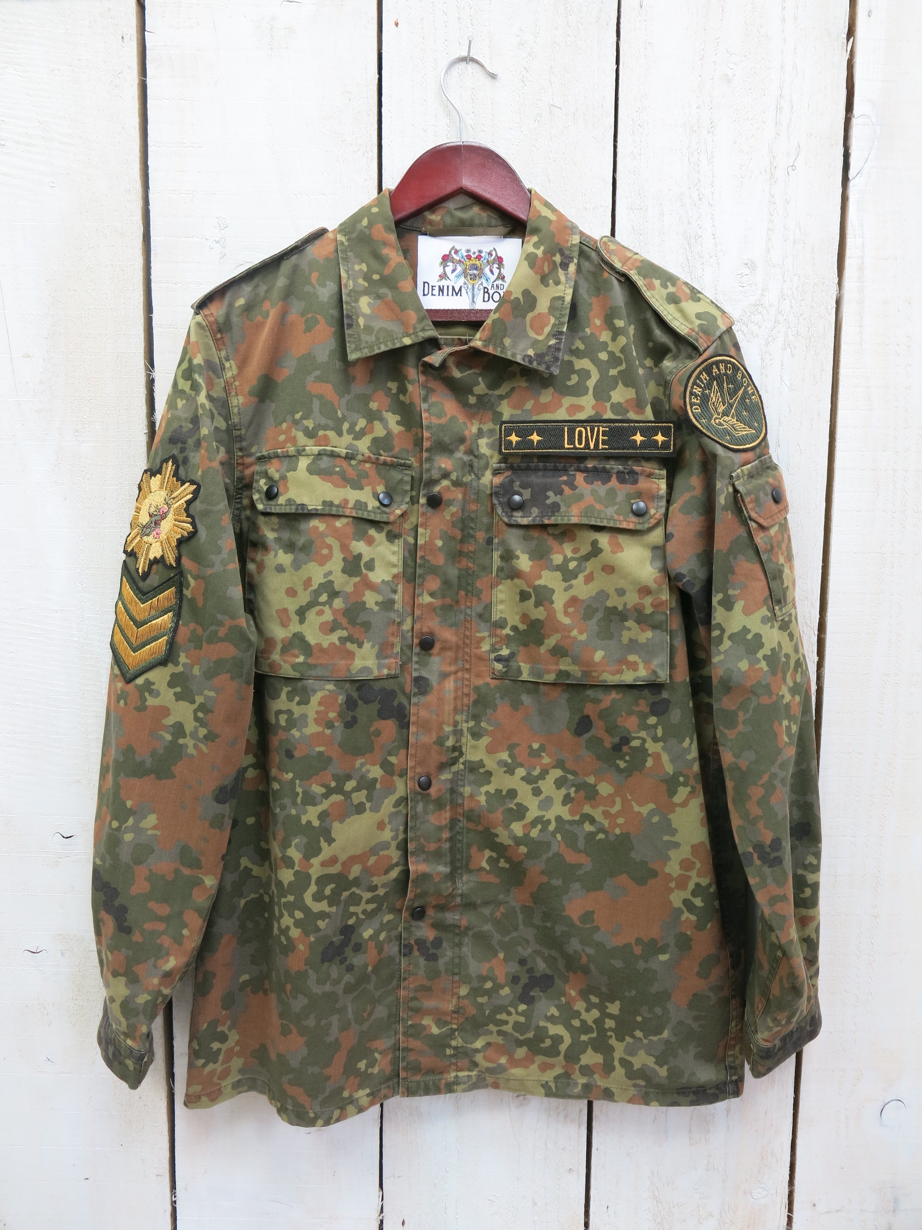 A camouflage military jacket with a "LOVE" embroidered chest patch and decorative military-style sleeve patches, shown from the front on a hanger.