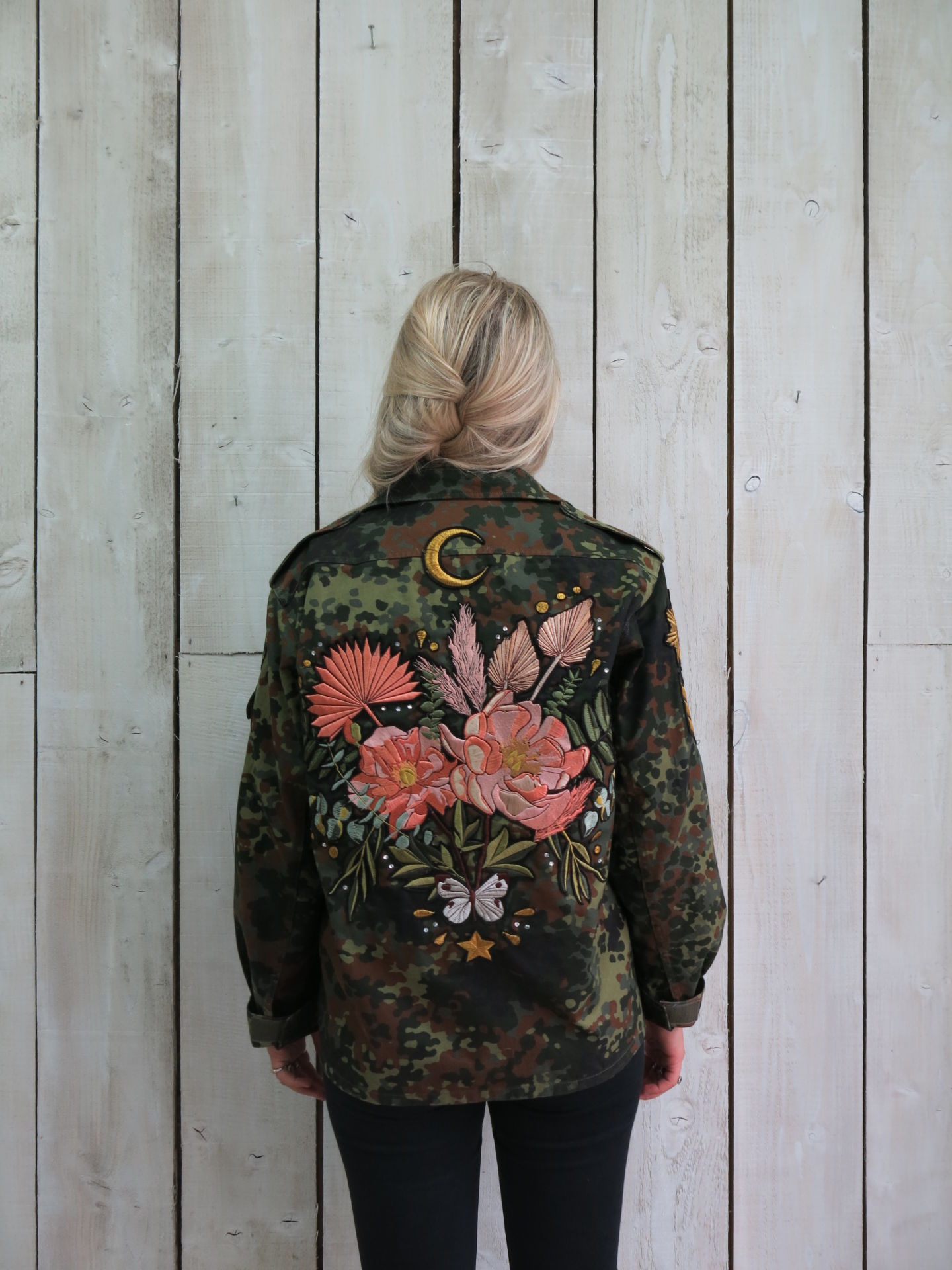 Person wearing a camouflage military jacket with a large embroidered floral bouquet, crescent moon, butterfly, and botanical design on the back.