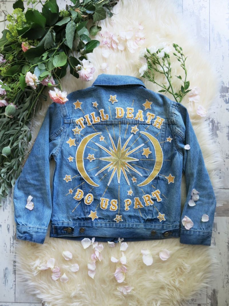 Embroidered denim jacket featuring a "Till Death Do Us Part" celestial design laid flat with flower petals.