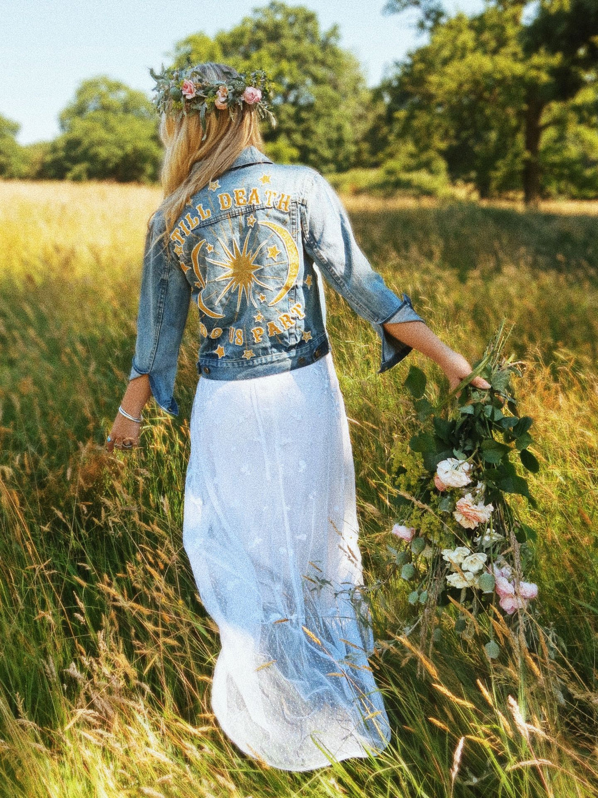 Woman walking through a meadow wearing an embroidered "Till Death Do Us Part" denim jacket and flower crown.