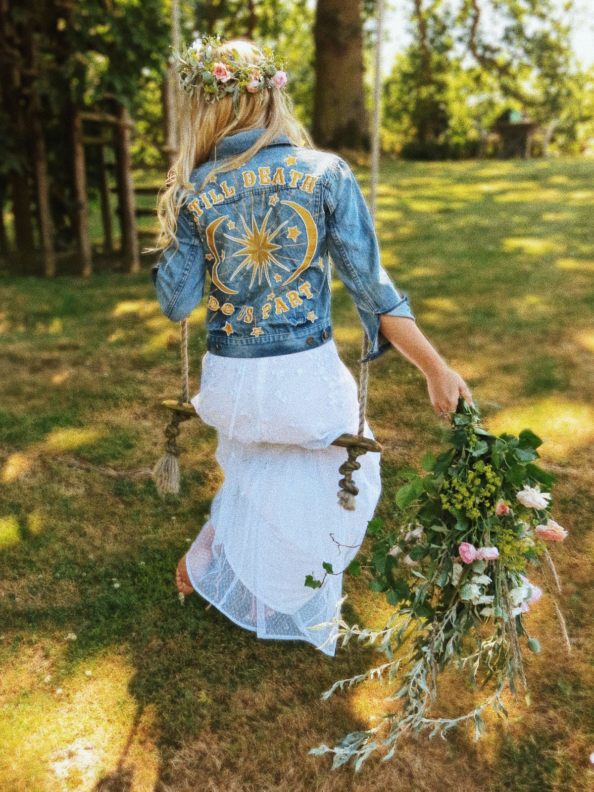 Bride wearing an embroidered "Till Death Do Us Part" denim jacket while holding a wildflower bouquet outdoors.