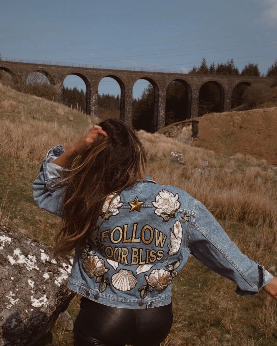 Light blue denim jacket with "Follow Your Bliss" embroidered back design, floral embroidery, and celestial motifs, shown from the back.