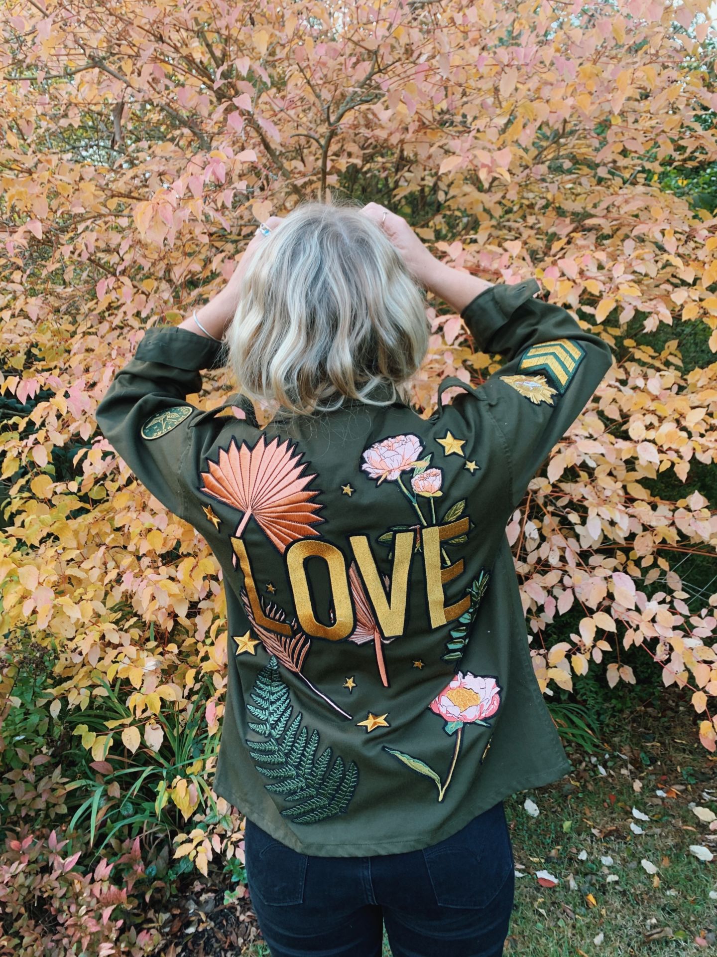 Olive green military jacket with floral embroidered back design and "LOVE" lettering, shown from the back.