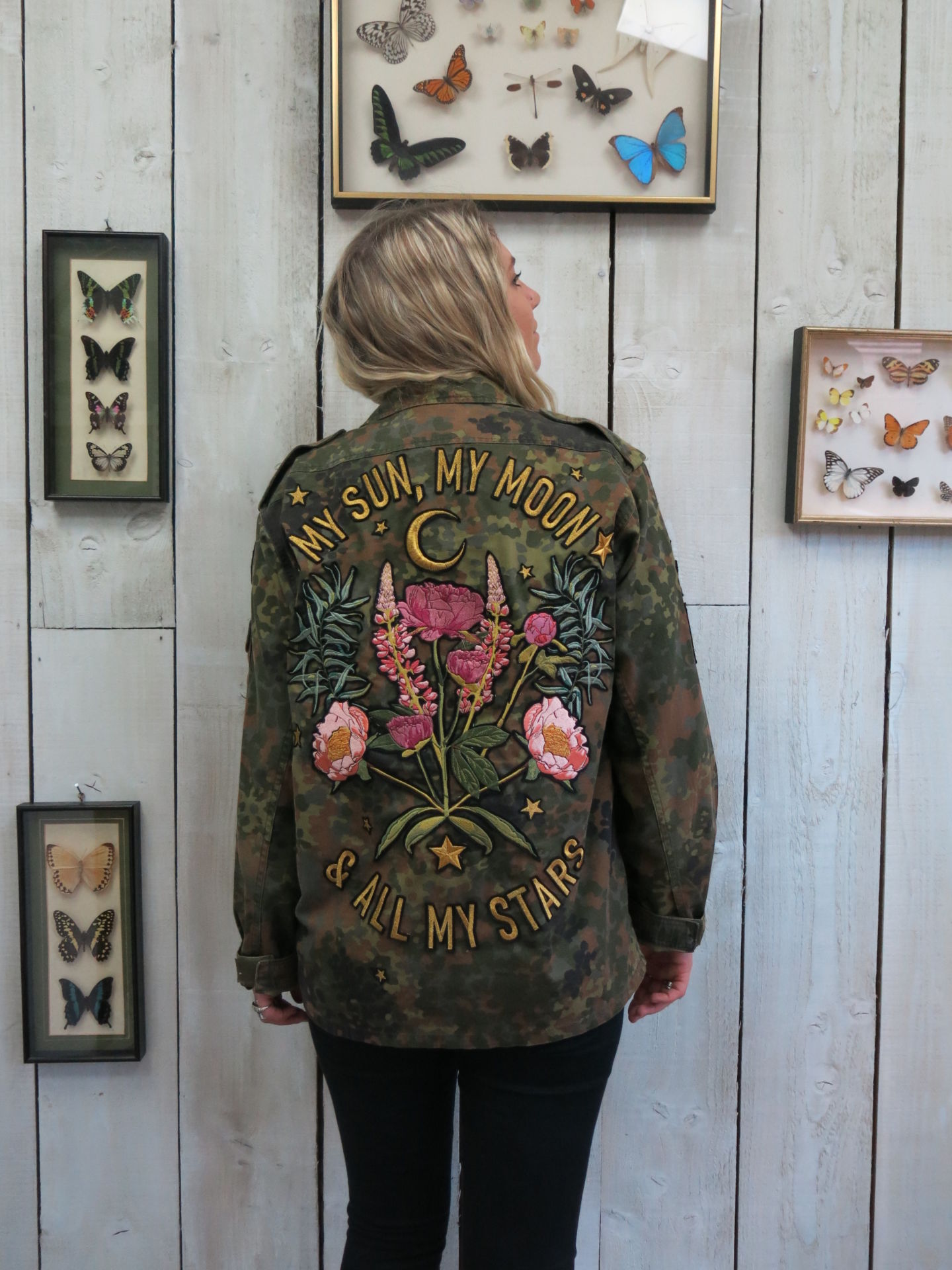 Back view of a person wearing a camouflage military jacket with embroidered floral bouquet, crescent moon, and "MY SUN, MY MOON & ALL MY STARS" lettering, standing against a light wood-paneled wall.