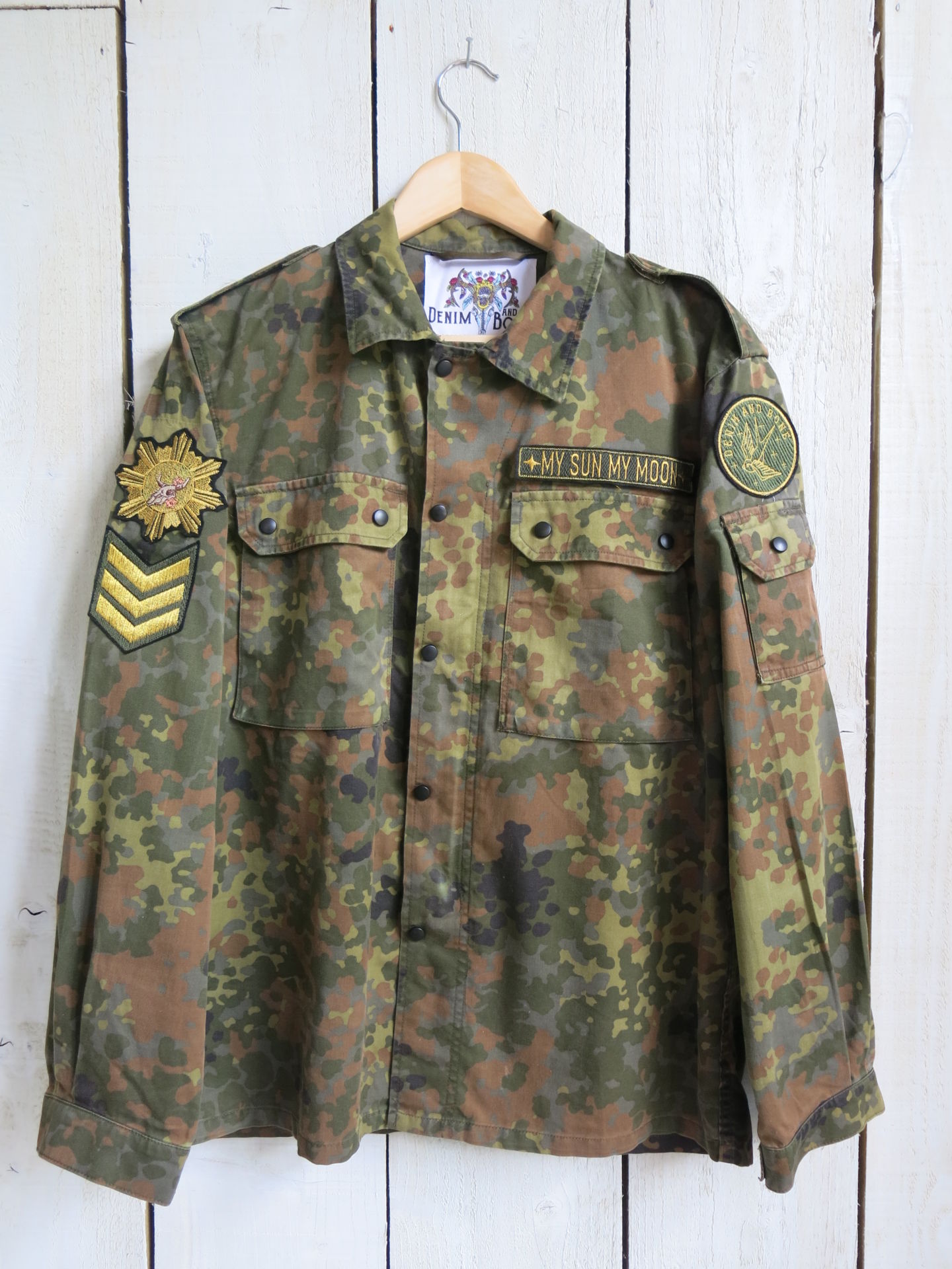 Front view of a camouflage military jacket with "MY SUN, MY MOON" chest patch, military-style sleeve patches, button-front closure, and flap chest pockets.