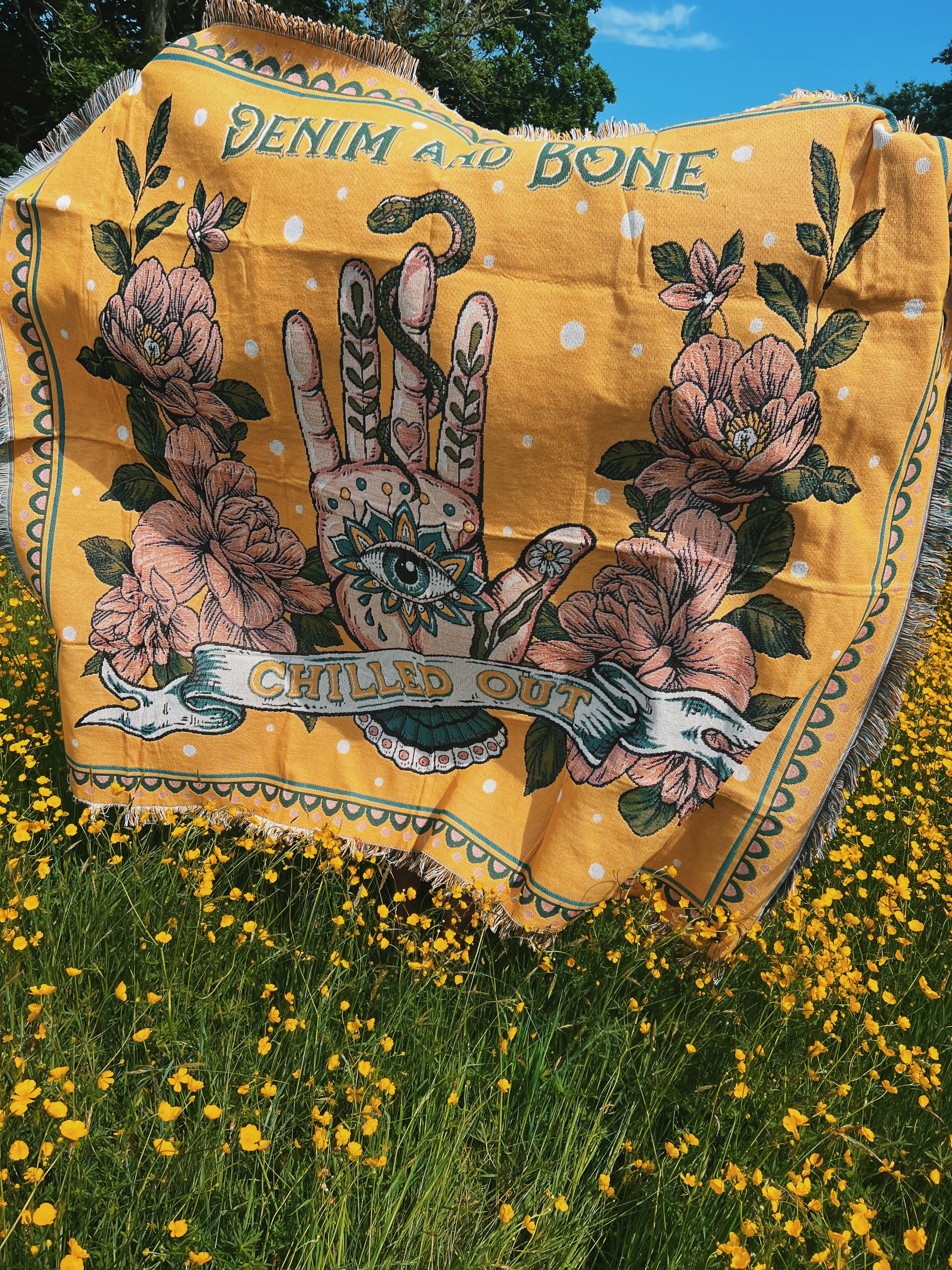 Yellow woven throw blanket with mystical hand, evil eye, pink flowers, and "Chilled Out" artwork laid on grass.