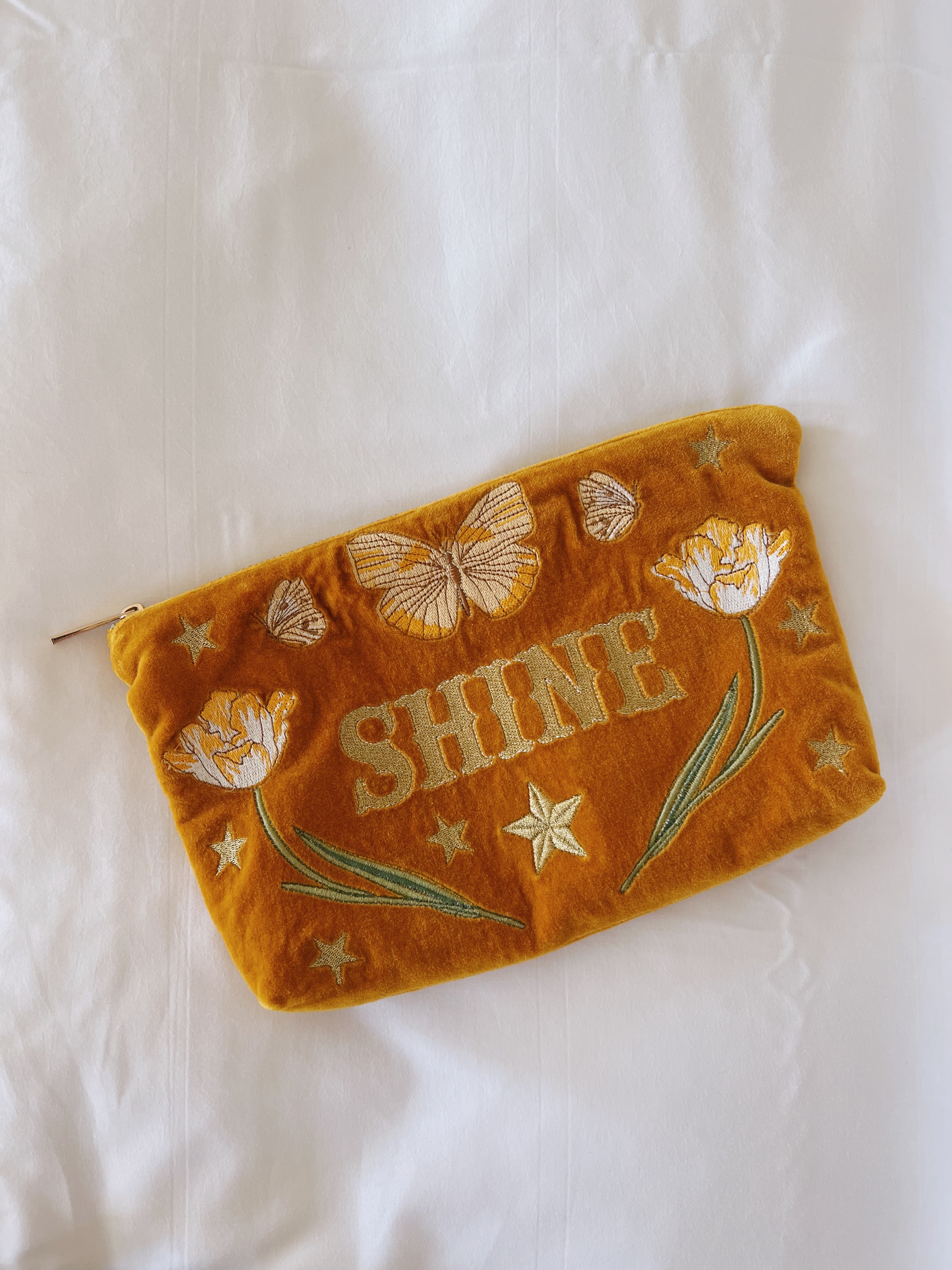 Golden yellow velvet zip pouch embroidered with "SHINE," butterflies, tulips, and stars on a white fabric background.