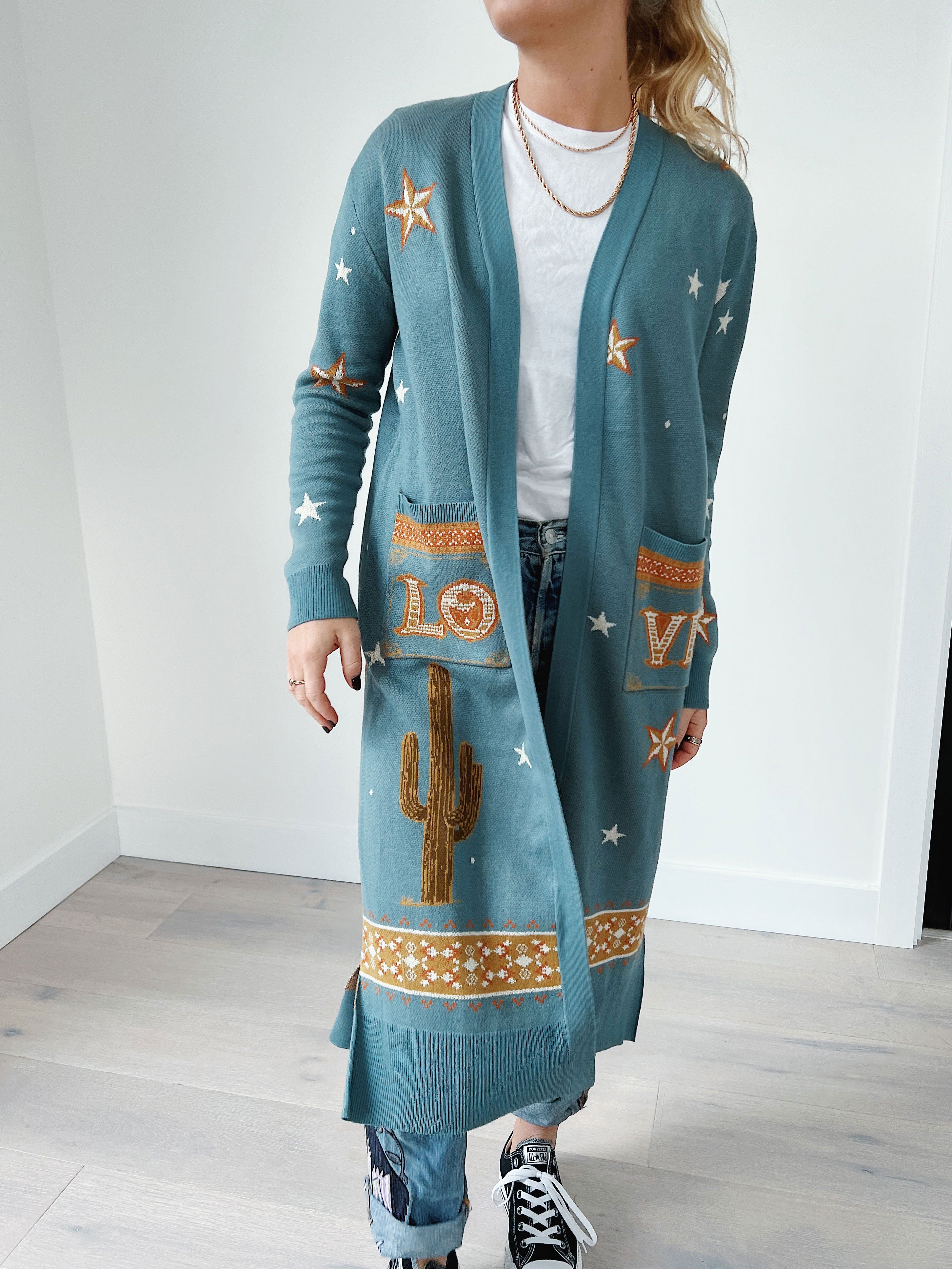 Front view of a person wearing a teal long knit cardigan featuring western-inspired knit designs with stars, cacti, geometric borders, and "LOVE" across the front pockets, layered over a white T-shirt and jeans.