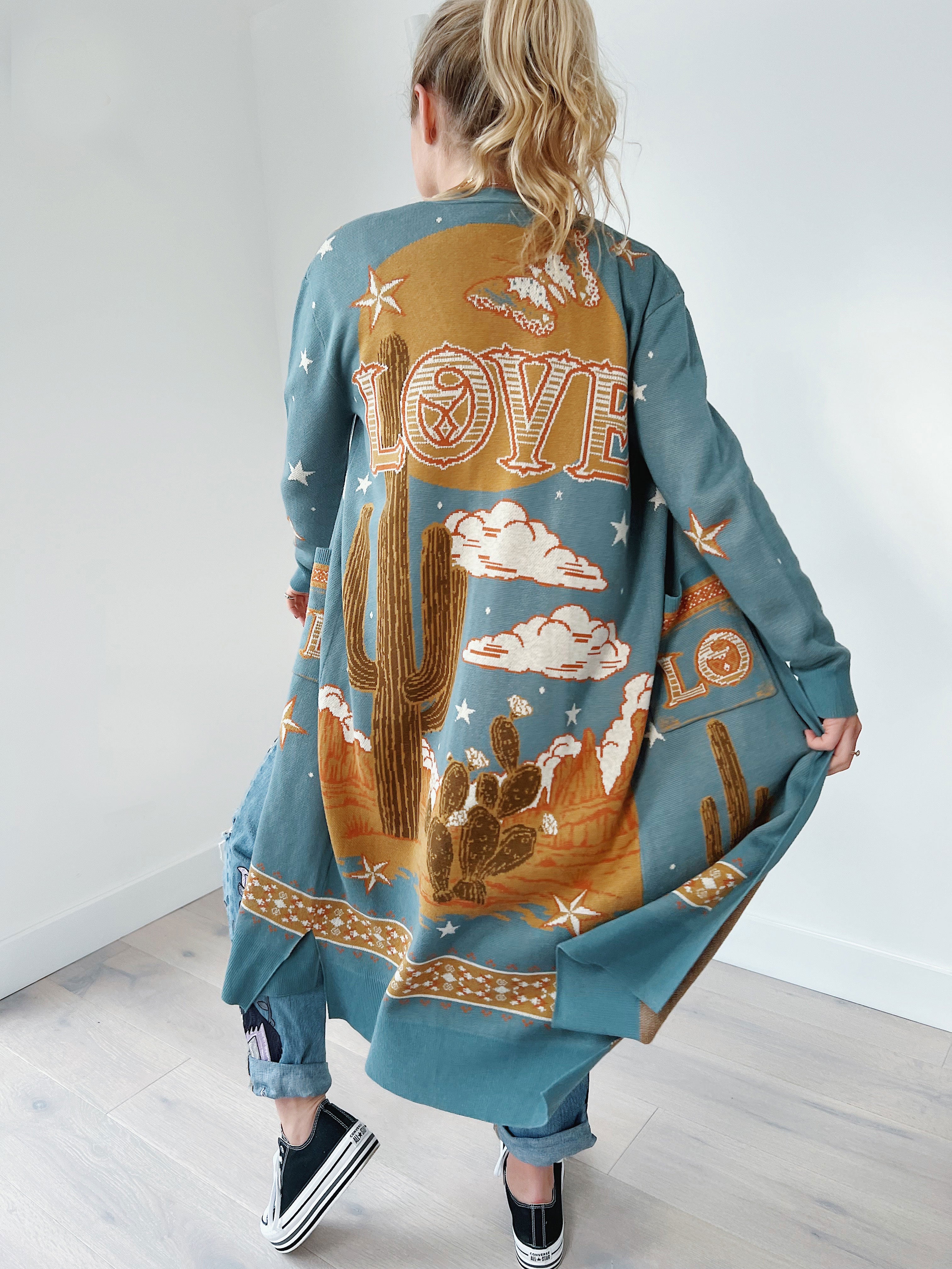 Back view of a person wearing a teal long knit cardigan featuring a western desert landscape knit design with a large cactus, stars, clouds, mesas, and the word "LOVE" on the back.