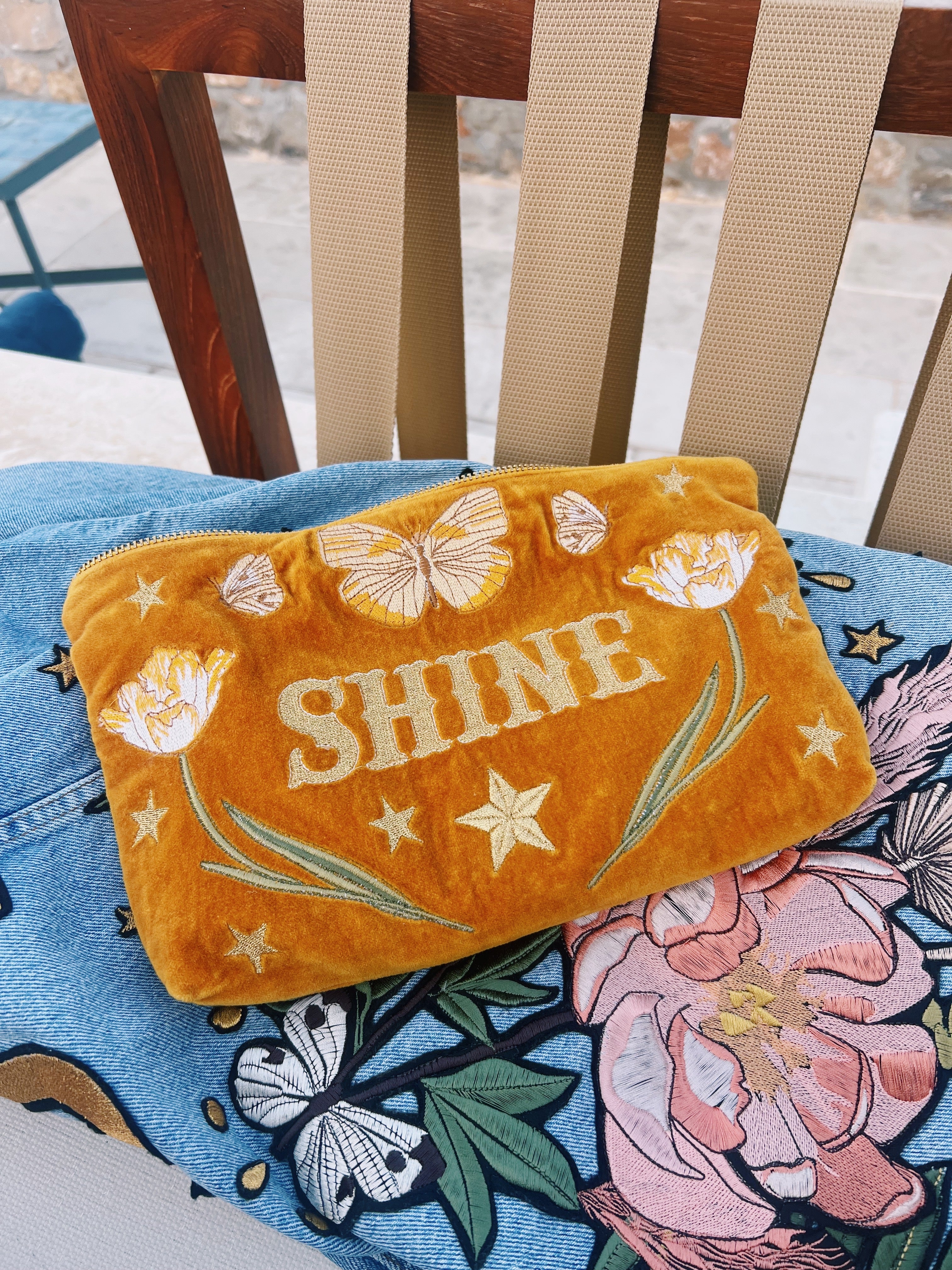 Golden yellow velvet zip pouch embroidered with "SHINE," butterflies, tulips, and stars, styled on a floral cushion.
