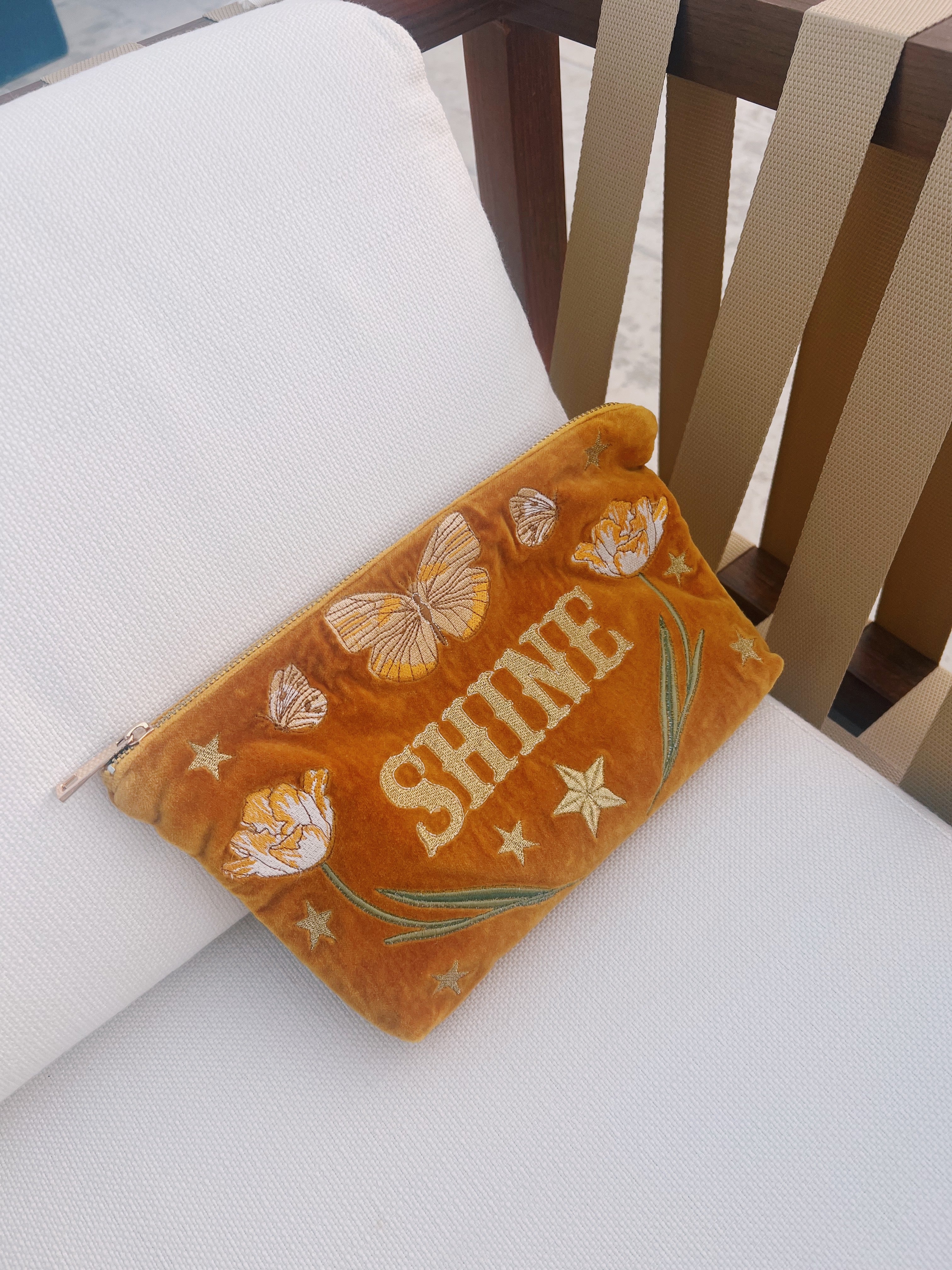 Golden yellow velvet zip pouch embroidered with the word "SHINE," butterflies, tulips, and stars, resting on a white chair.