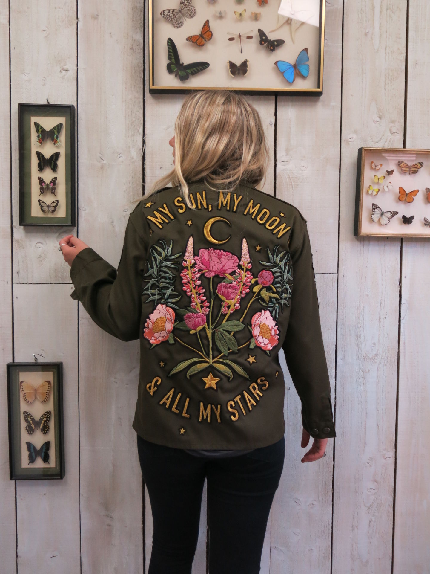 Back view of a person wearing an olive green military jacket with embroidered floral bouquet, crescent moon, and "MY SUN, MY MOON & ALL MY STARS" lettering, standing against a light wood-paneled wall.