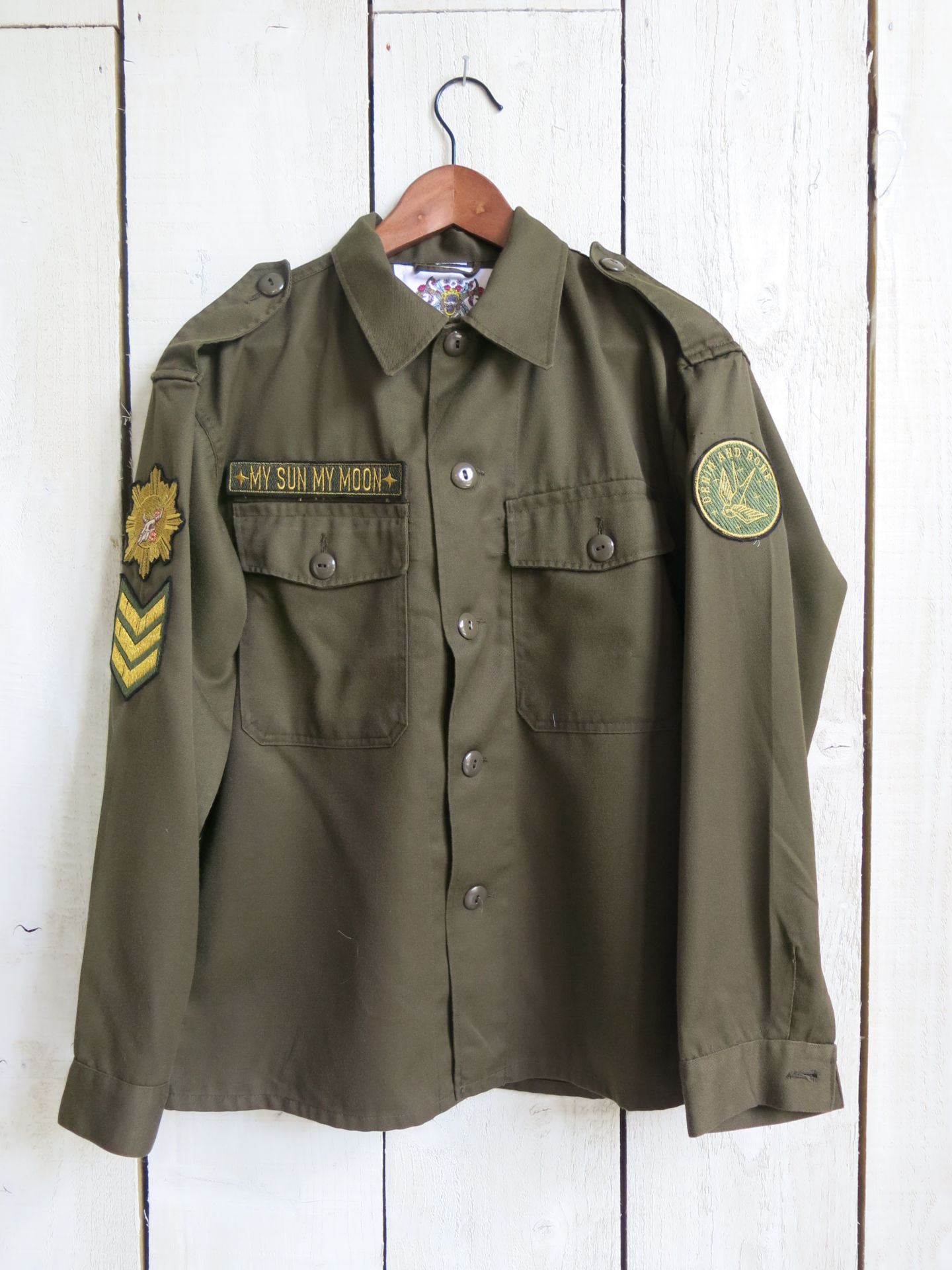 Front view of an olive green military jacket with "MY SUN MY MOON" embroidered chest patch, decorative sleeve patches, and button-front design.