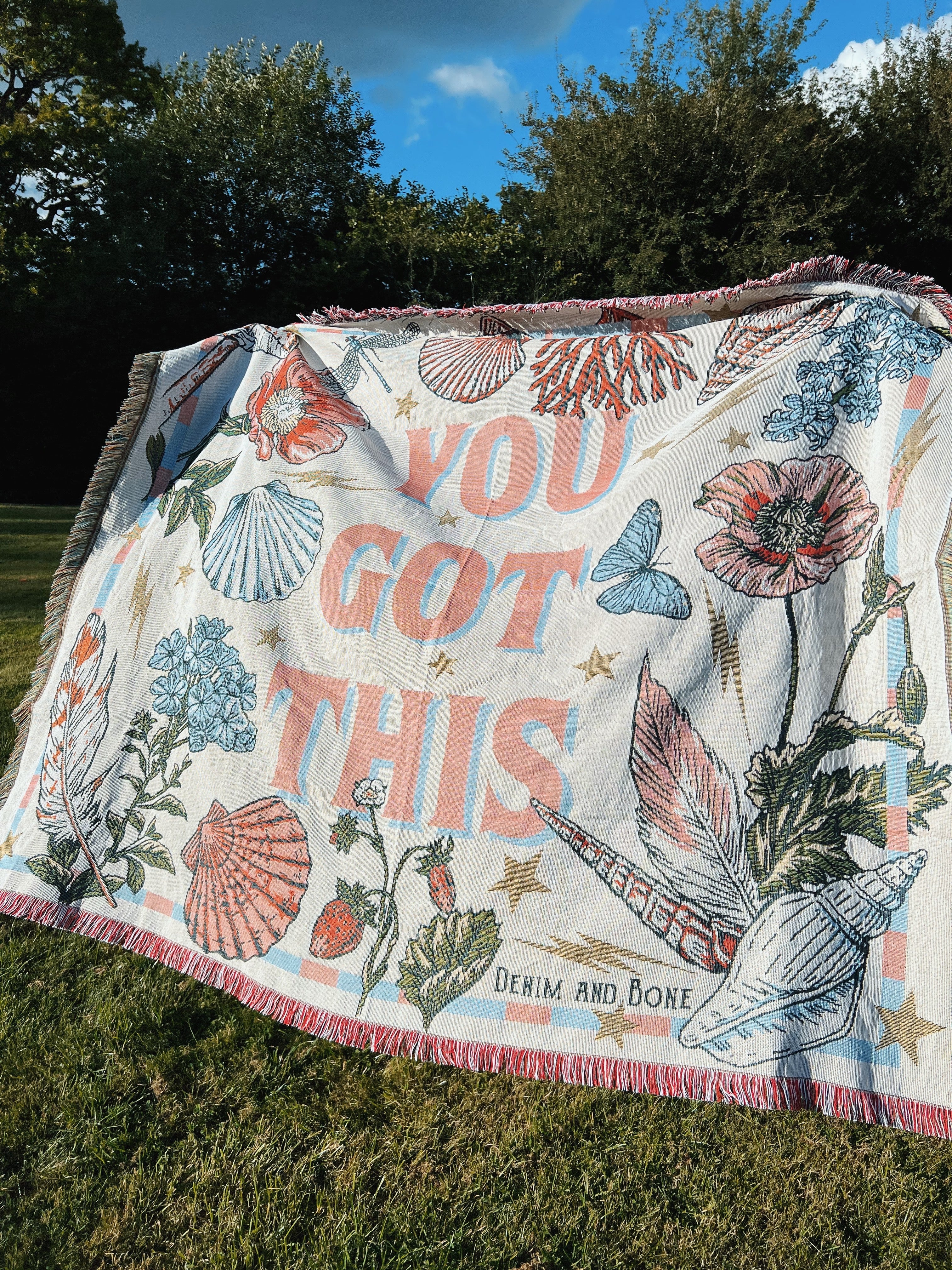 "You Got This" woven throw blanket with wildflowers, seashells, and butterflies displayed outdoors on the lawn.