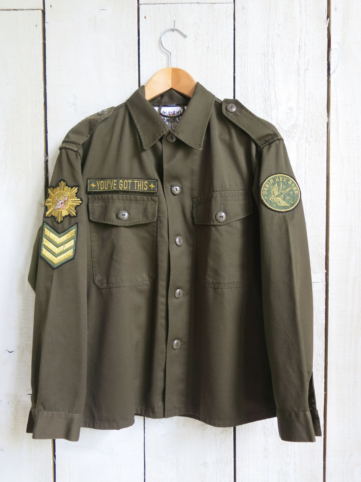 Front view of an olive green military jacket with "YOU'VE GOT THIS" chest patch, military-style sleeve patches, button-front closure, and flap chest pockets.